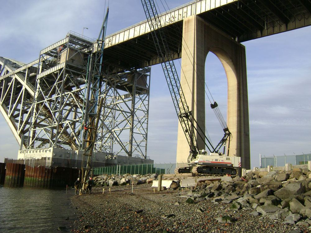 Crawler crane working on the shoreline beneath a large steel arch bridge
